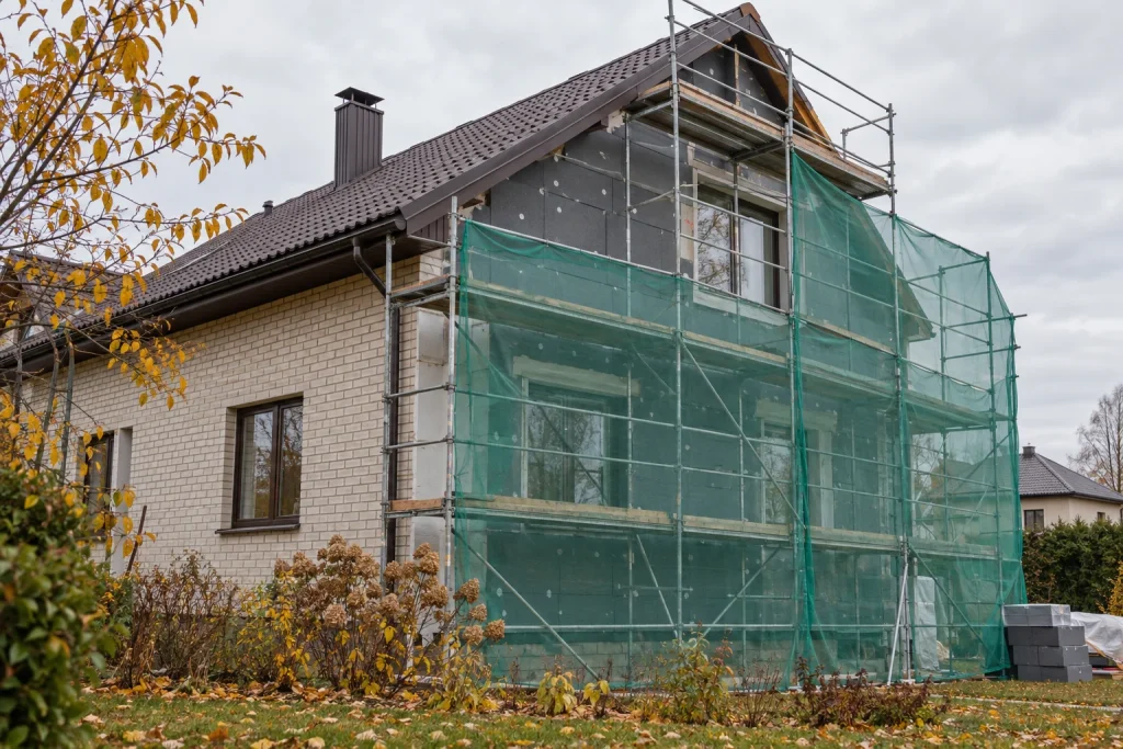 Šiltinamo individualaus namo fasadas su pastoliais ir apsauginiu tinklu rudenį